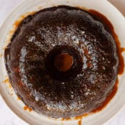 Bundt tin-shaped glossy sticky date pudding on a white platter in a puddle of butterscotch sauce.