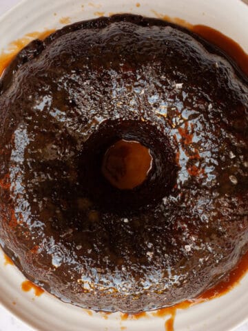 Bundt tin-shaped glossy sticky date pudding on a white platter in a puddle of butterscotch sauce.