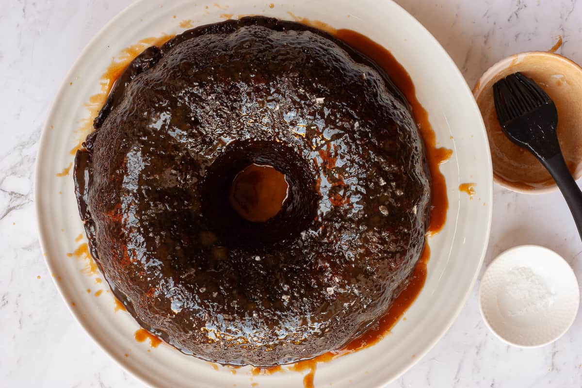 Bundt tin-shaped glossy sticky date pudding on a white platter in a puddle of butterscotch sauce.