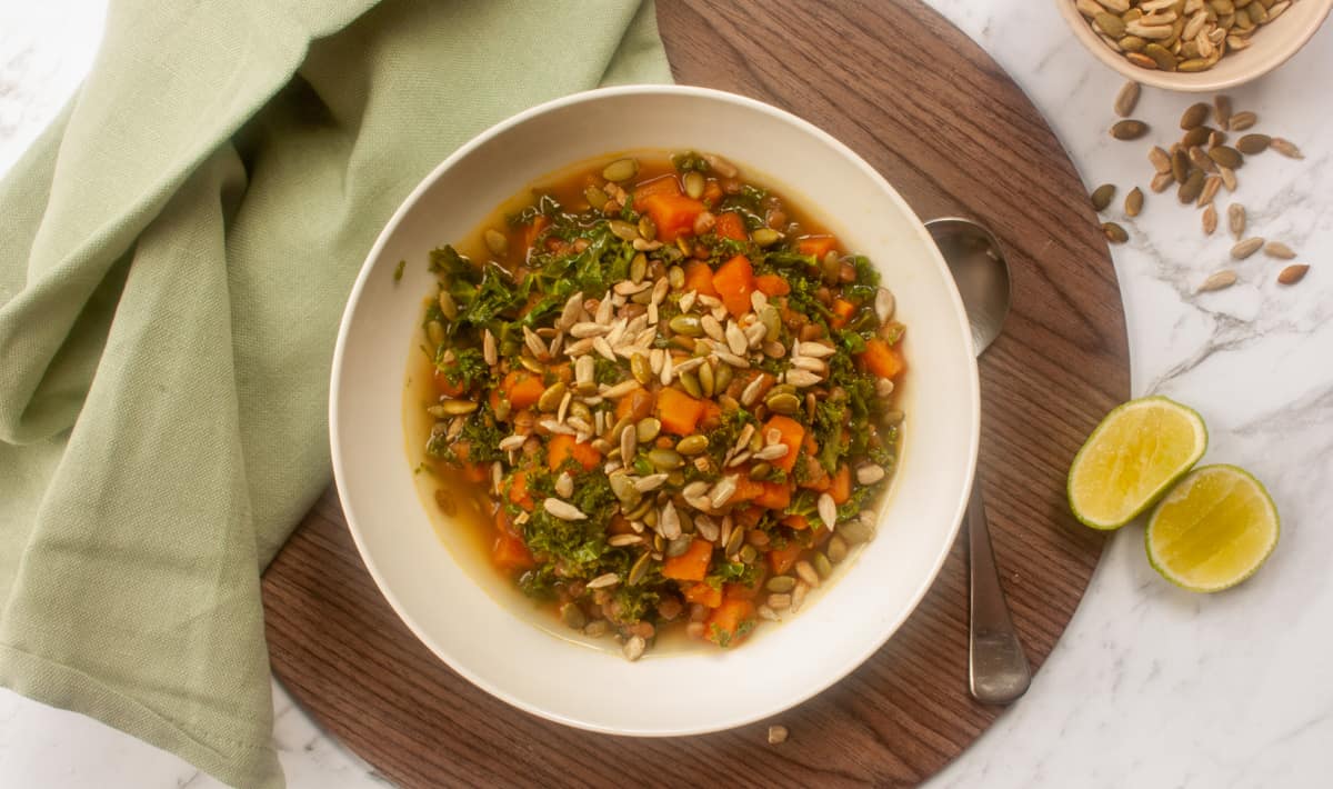 Overhead shot of stew in a white bowl set on a circular wooden placemat. Some lime cheeks and toasted seeds to the side.