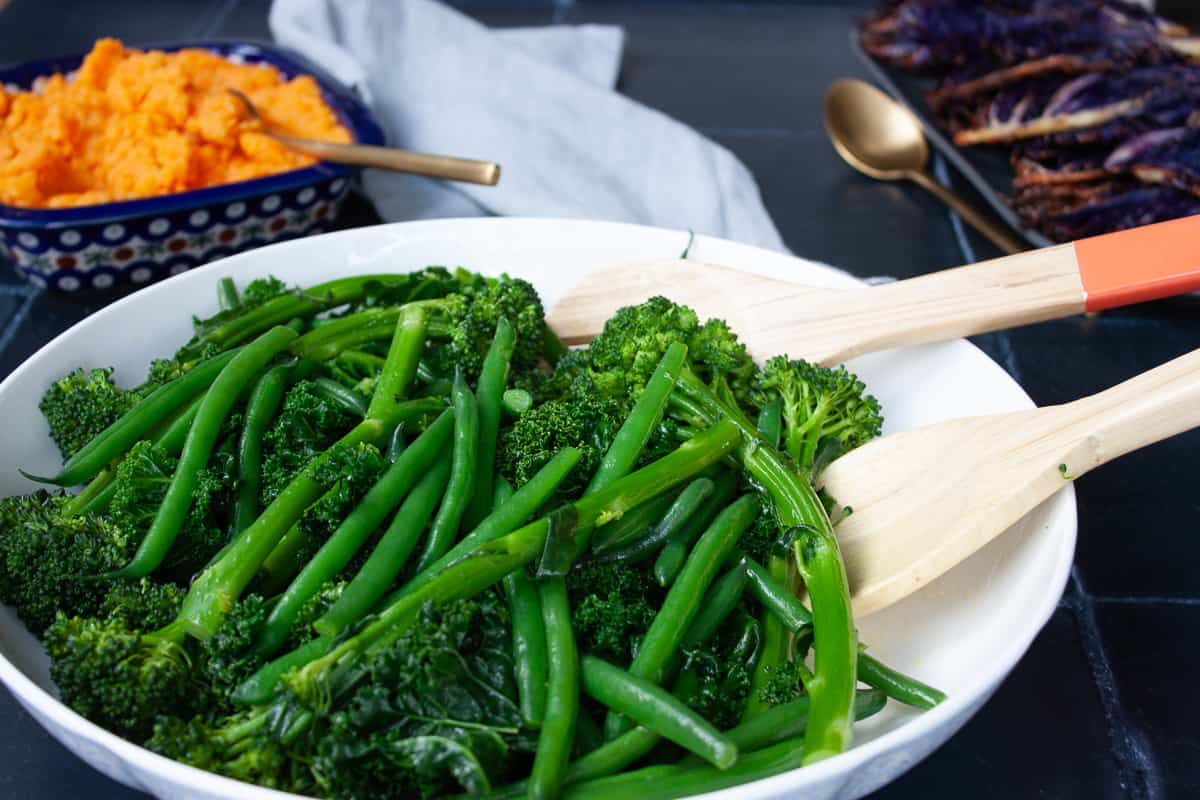 Tender-crisp green greens in a white bowl with other vegetable side dishes in the background.