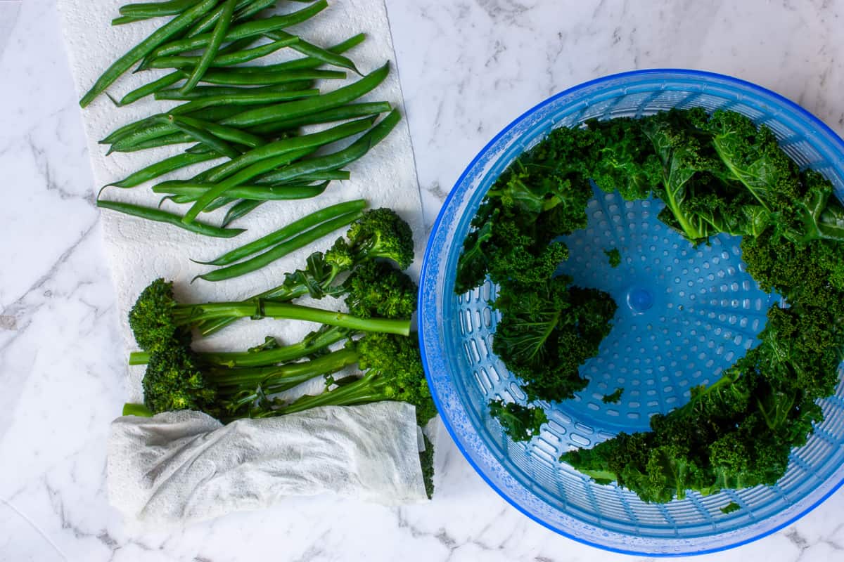 Kale leaves spun dry against the edges of a salad spinner. Broccolini partially rolled up in paper towel.